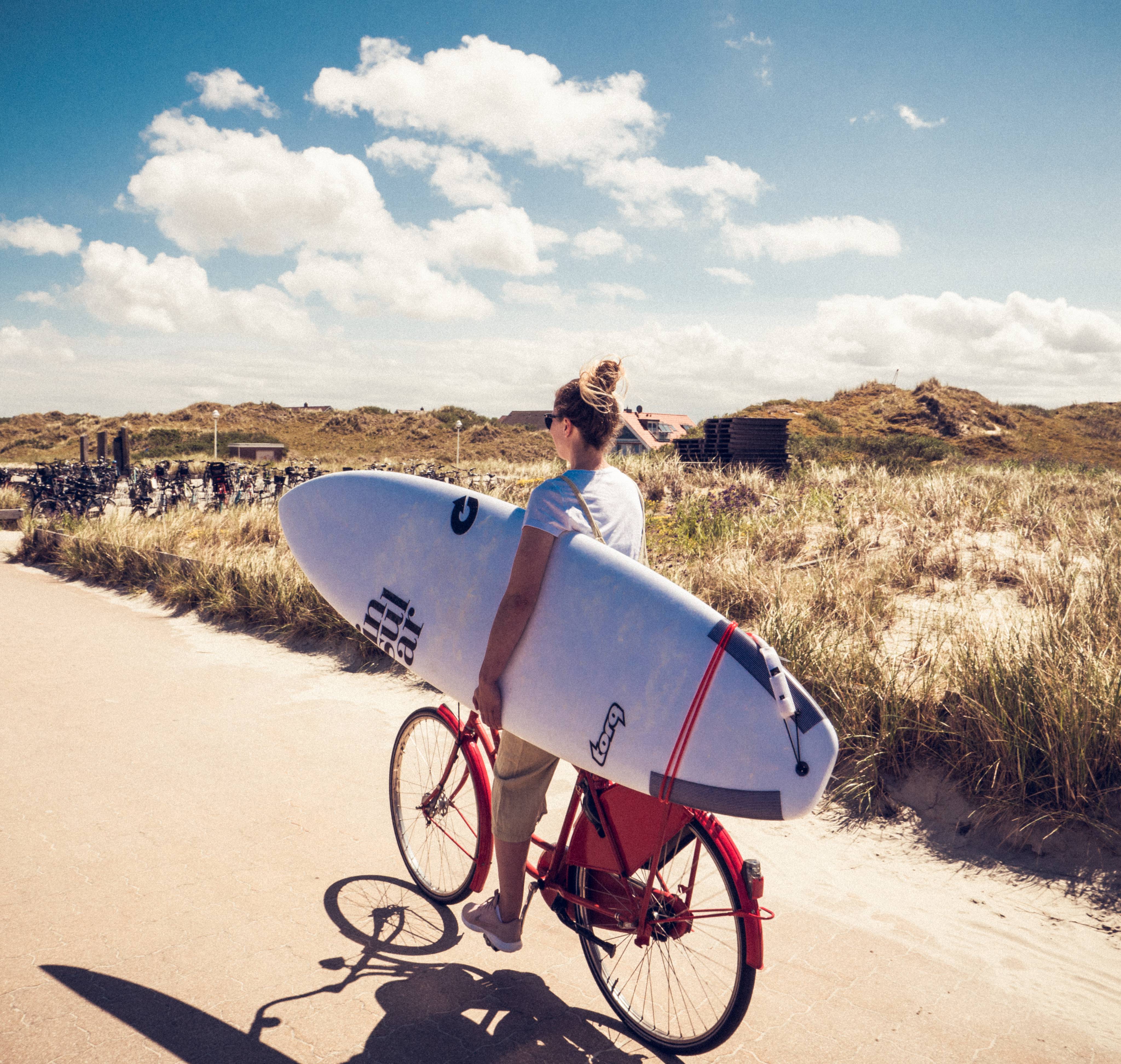 Frau fährt auf dem Rad mit Surfbrett unter dem Arm druch die Dünen